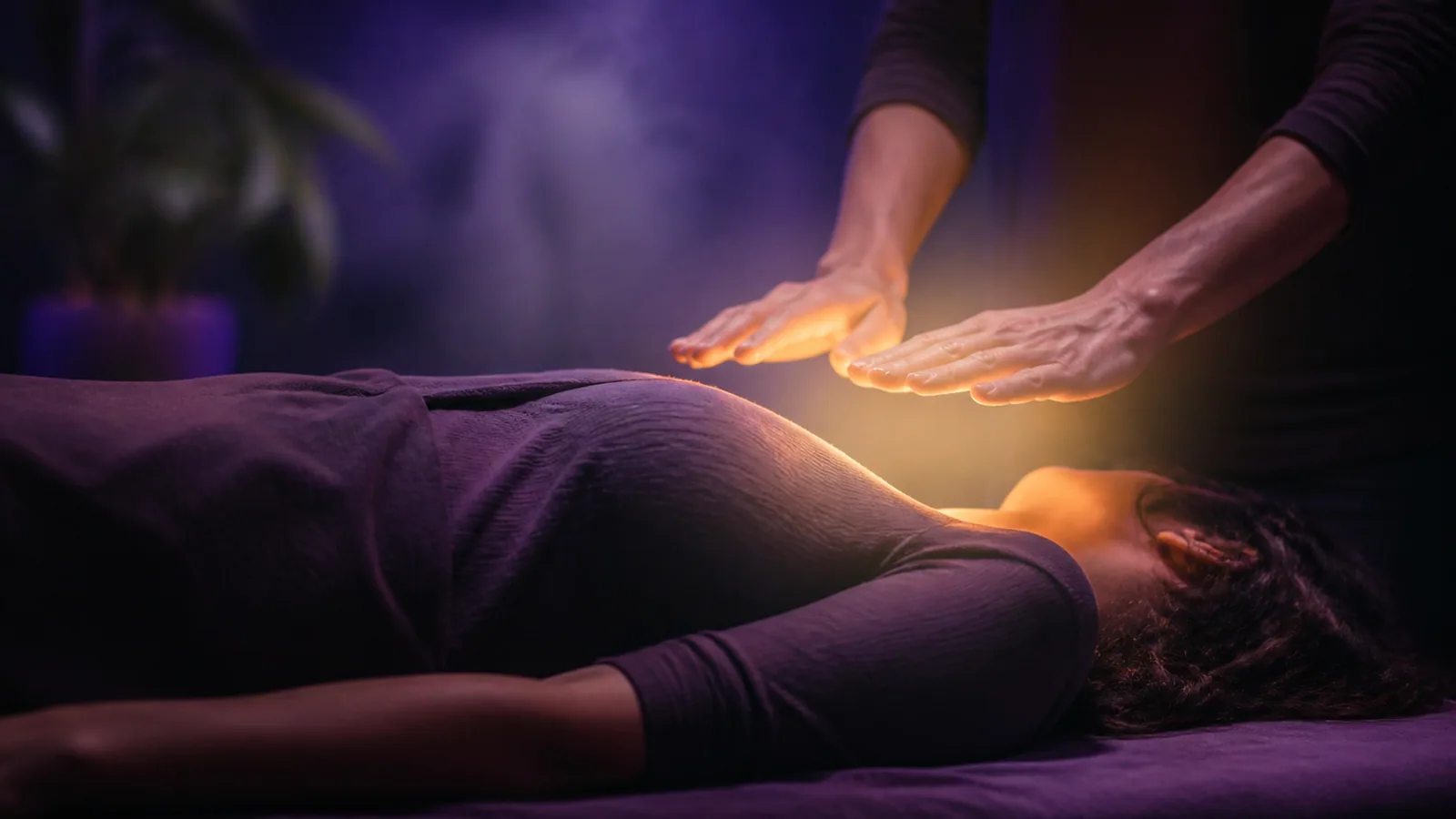 Mains de praticien légèrement levées au-dessus du buste d'une personne allongée sur une table, lumière dorée tamisée et fond violet, séance Reiki Shamballa